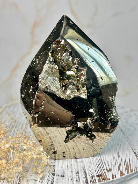 Golden pyrite flame crystal polished into flame shape, shimmering metallic surface displayed on a distressed wooden board with cream dried flowers.