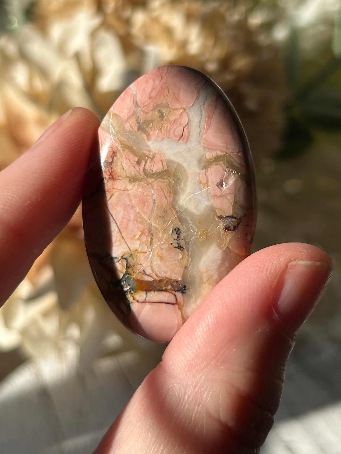 A polished oval stone with pink, cream, and brown marbled patterns, held between fingers in bright natural light.