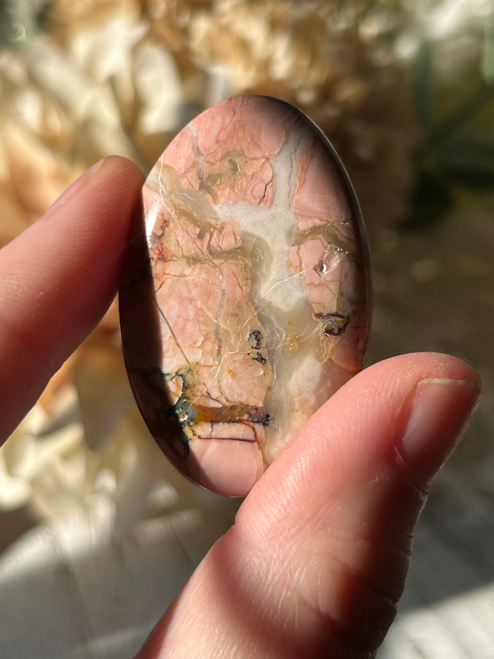 A polished oval stone with pink, cream, and brown marbled patterns, held between fingers in bright natural light.