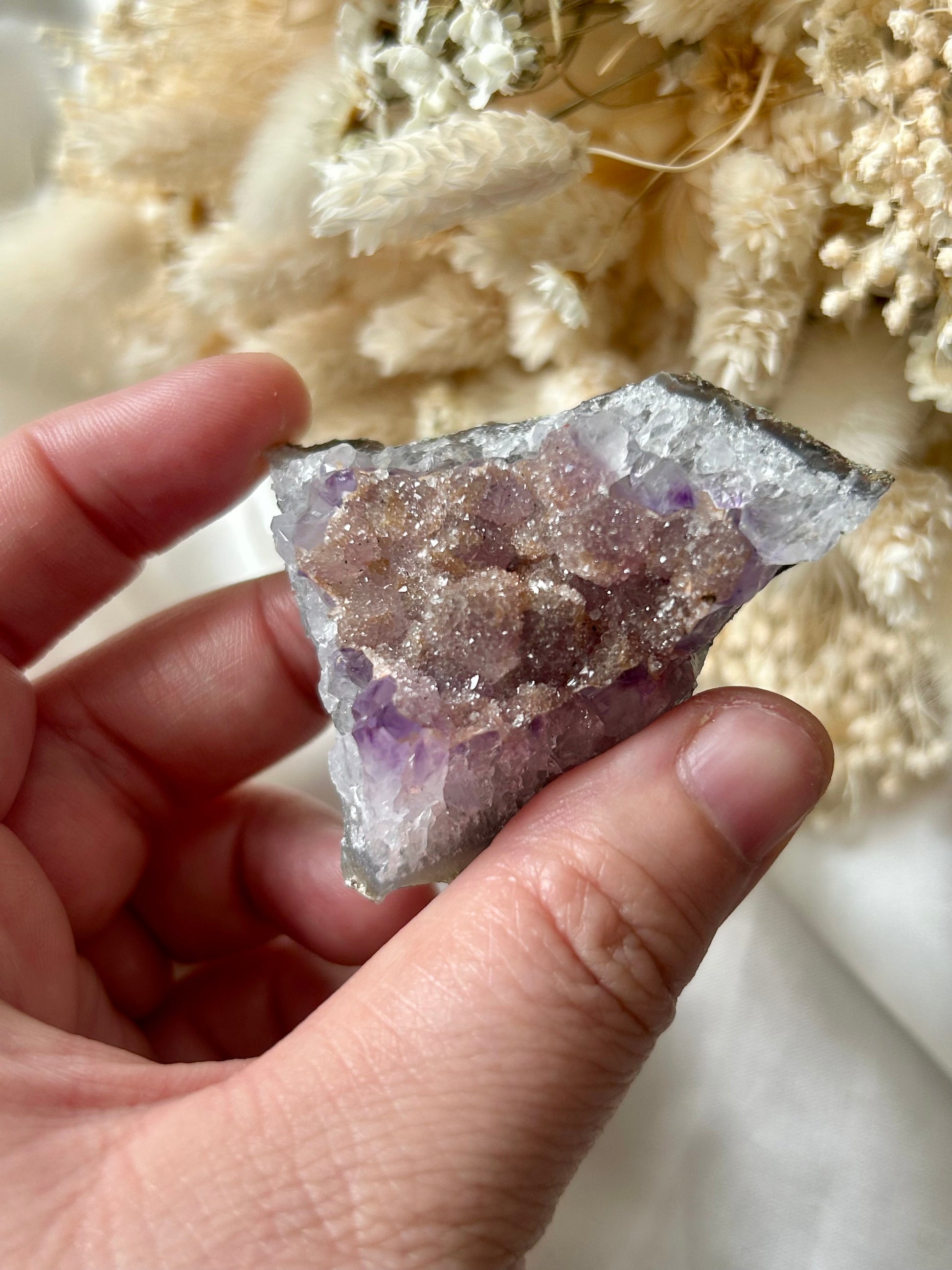 Small druzy rainbow sugar geode clusters with pastel tones and glittering crystal surfaces, displayed on ivory silk with cream dried flowers in the background.