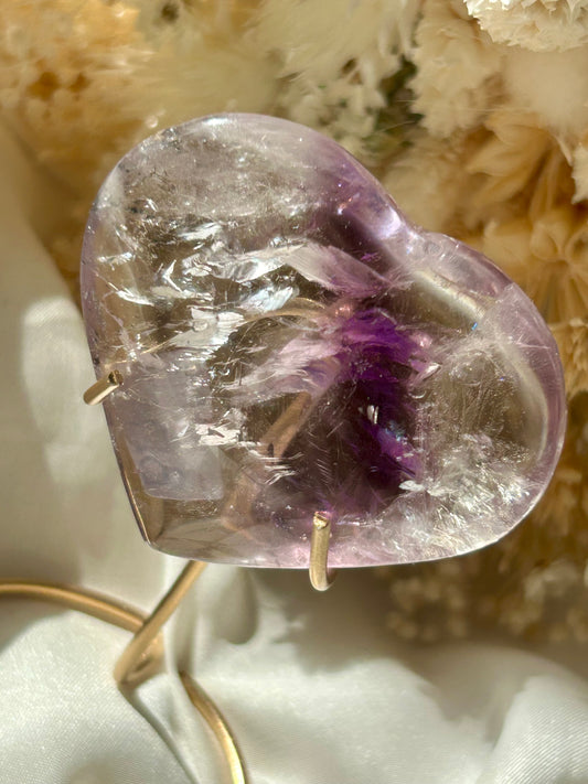 Polished Amethyst heart crystal from Brazil with a deep purple phantom inclusion and visible rainbow flashes, resting on ivory satin with dried flowers in the background