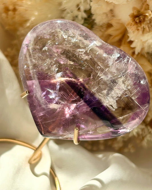 Polished Amethyst heart crystal from Brazil with a deep purple phantom inclusion and visible rainbow flashes, resting on ivory satin with dried flowers in the background
