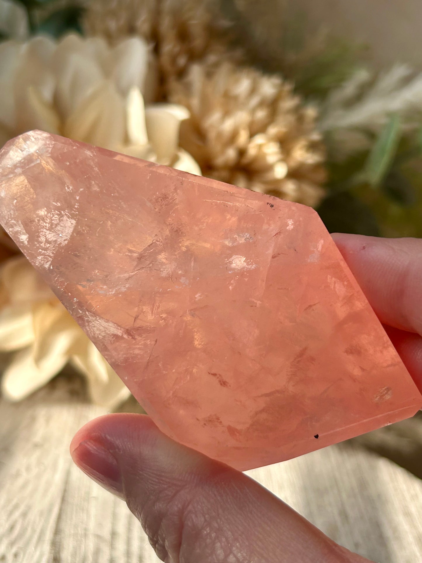 A hand holding a rough, faceted peach-pink crystal against a soft-focus background of dried flowers and light wood.