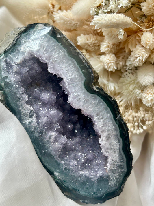 Purple and lavender sugar geode with sparkling druzy crystals and deep navy-violet core, displayed on ivory silk with soft cream dried flowers in the background.

