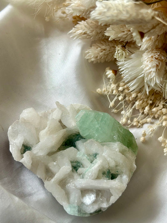Green Apophyllite and Stilbite crystal cluster with pale green translucent points and peach-toned blades, displayed on ivory silk with soft dried flowers in the background.
