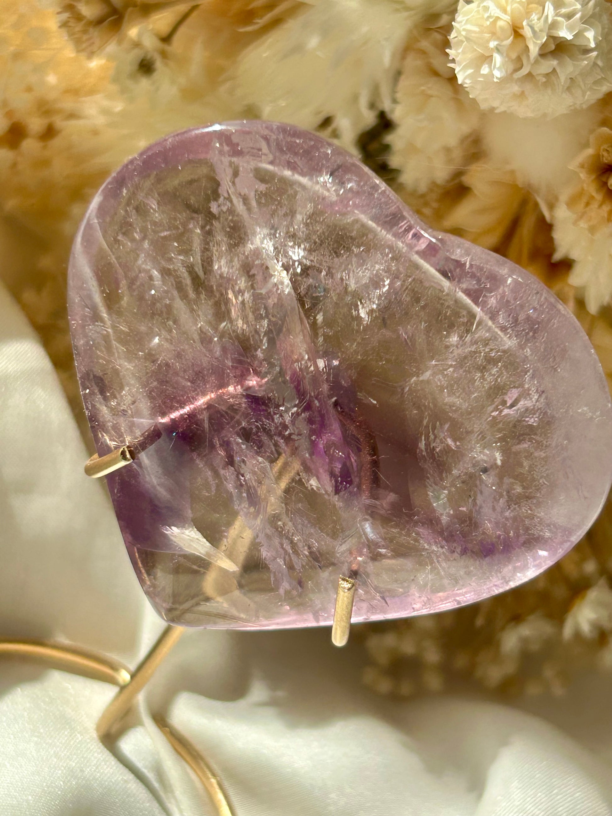 Polished Amethyst heart crystal from Brazil with a deep purple phantom inclusion and visible rainbow flashes, resting on ivory satin with dried flowers in the background