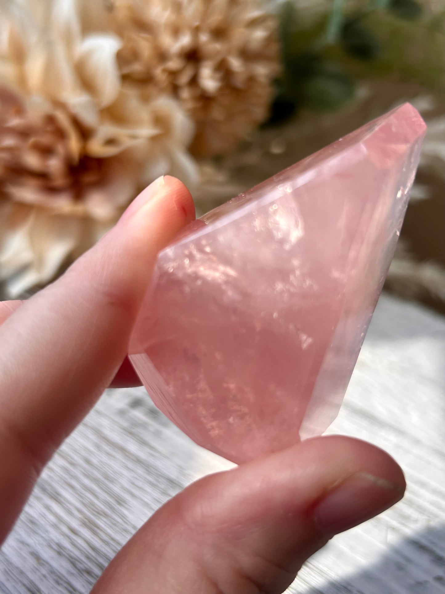 A hand holding a polished pink rose quartz crystal point against a soft-focus background of dried flowers and a light wooden surface.