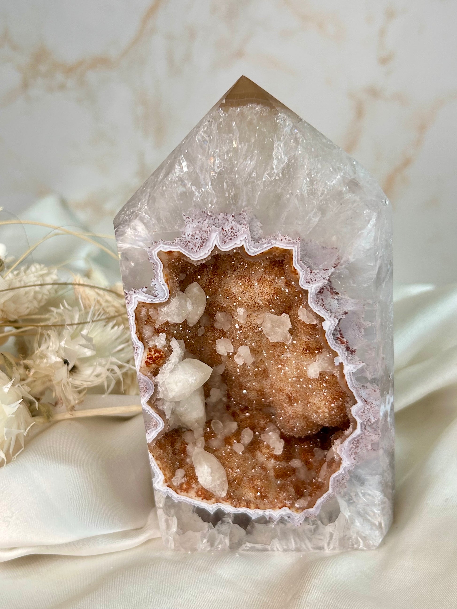 Brazilian Agate tower featuring a warm caramel sugar-druzy cavity with sparkling crystals, soft peach and white banding, and natural formations. Displayed against neutral dried florals and a cream fabric backdrop.