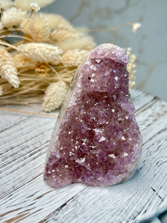 Amethyst cut base from Uruguay with vibrant violet crystal points and polished edges, displayed on a rustic white stand beside dried florals.