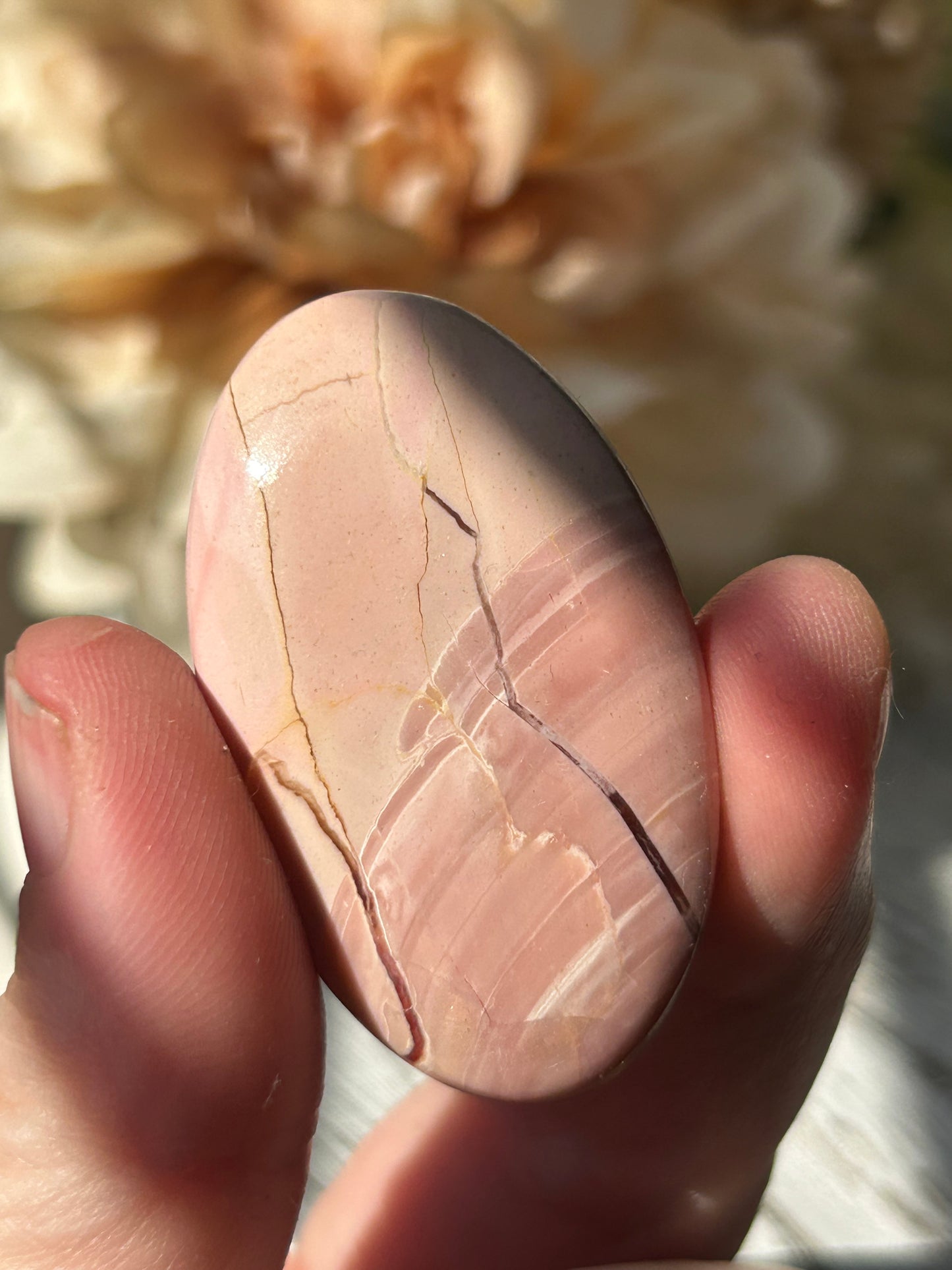 A hand holding a polished oval pink stone with natural cracks and veining, photographed in bright natural light against a blurred beige background.