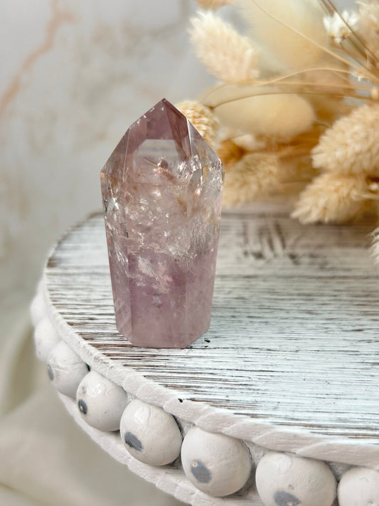 UV-reactive Amethyst tower with internal rainbows and vivid inclusions, polished point displayed on a rustic white wooden stand with dried flowers in the background